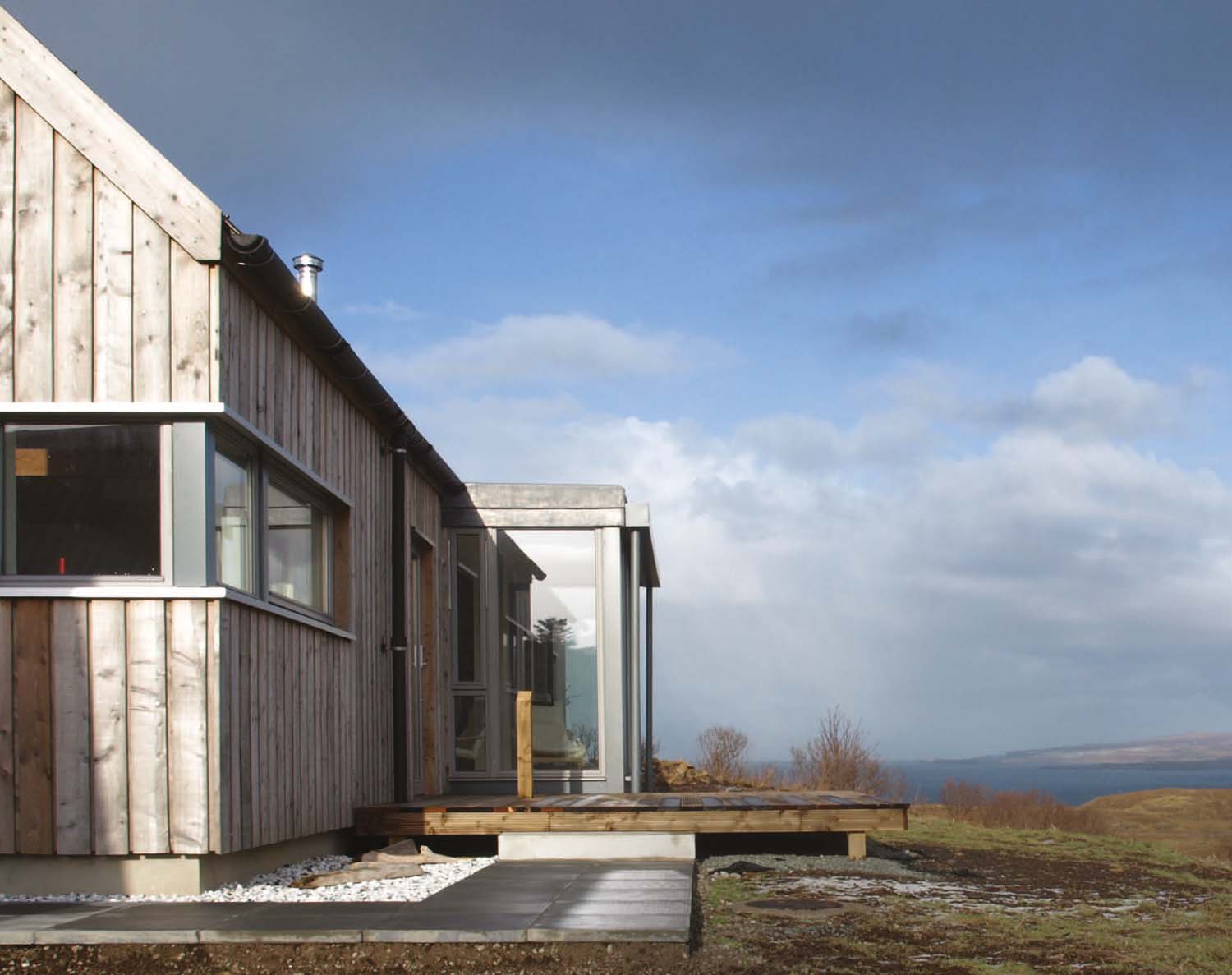 The outdoor decking and an extension at the Long House overlooking the sea and its rural landscape. Large windows frame the extension's walls.