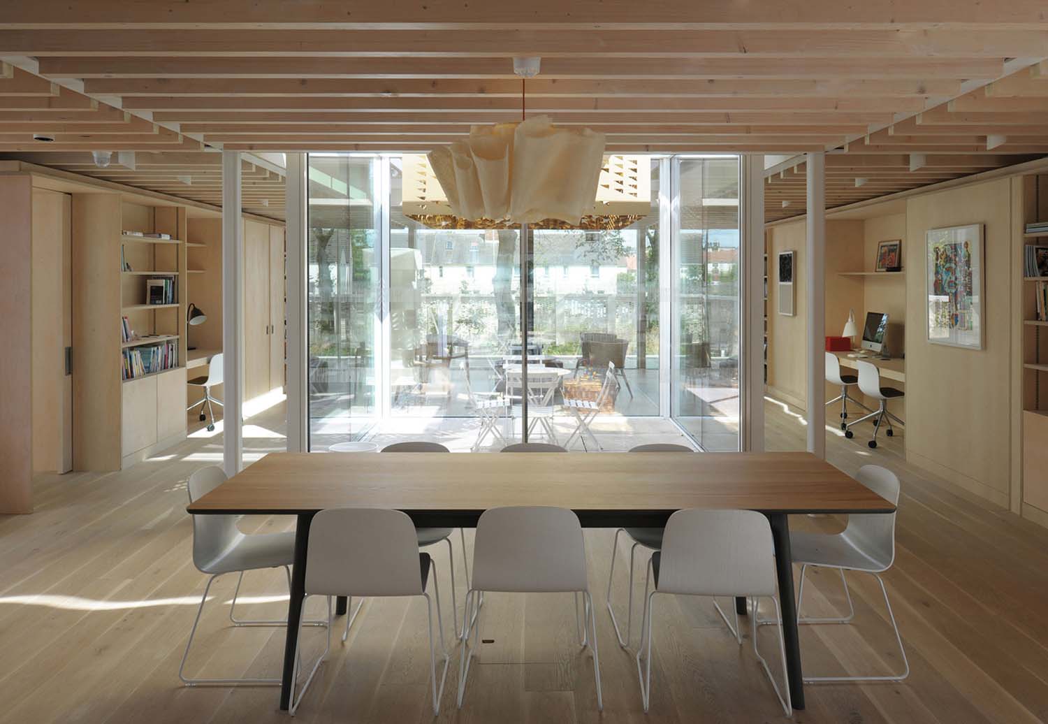 An interior view of a large social space with blonde wooden walls and furniture looking out towards a window leading to a green space