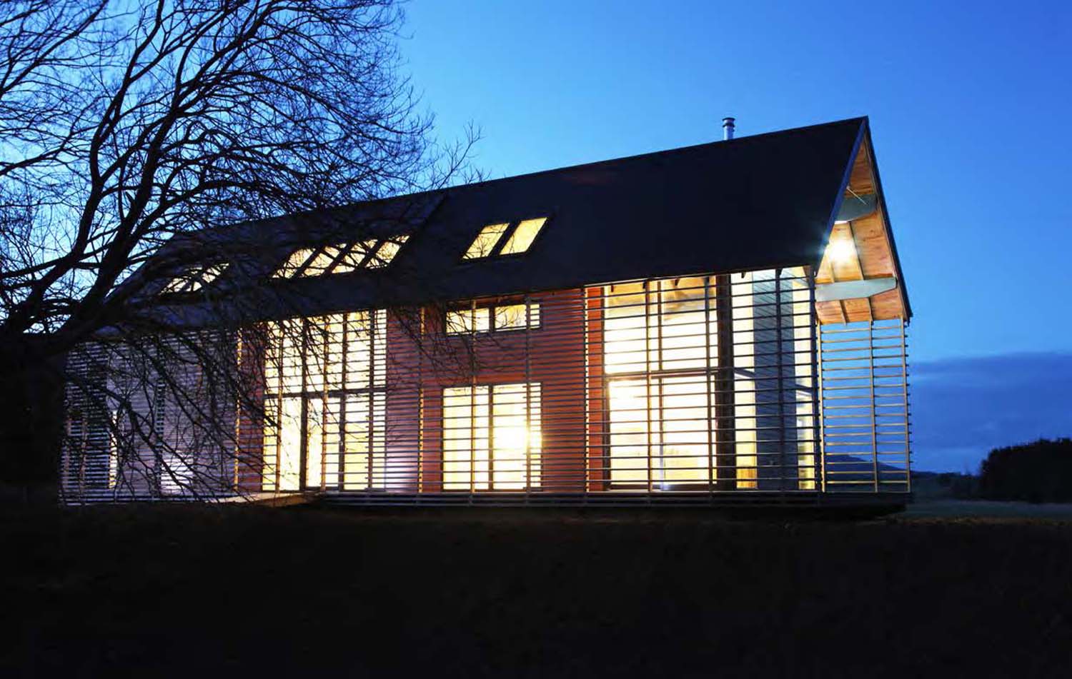 A photograph of  large timber building at nighttime showing light coming from the house which is silhouetted against a blue night sky