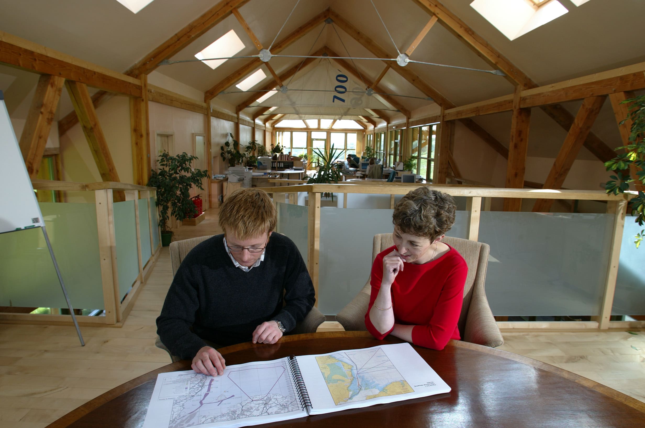 Two people working on architectural drawings on the first floor of an office. The interiors are long and narrow with skylights. The timber frame of the building is visible.