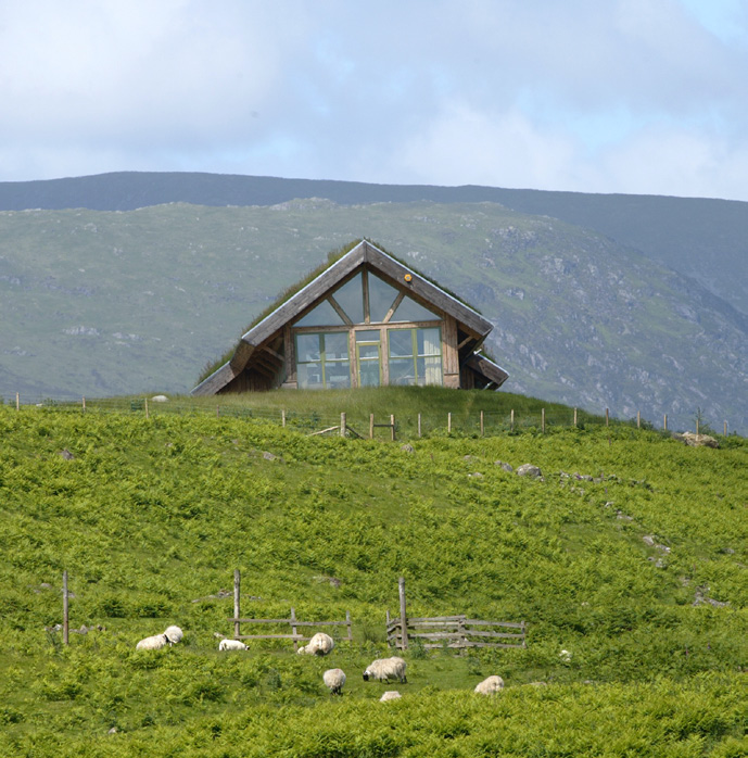 A rear view of a small building nestled on a hill. There are mountains in the background and sheep grazing below the hill.