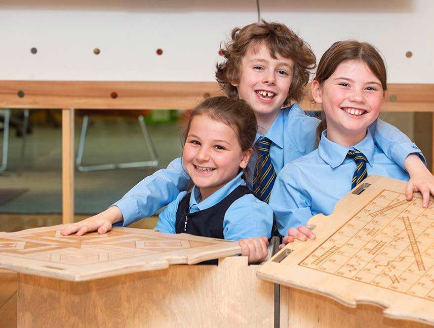 Three primary school pupils dressed in uniforms smile at the camera. They are holding pieces of custom made wooden furniture for their school, that they helped design.