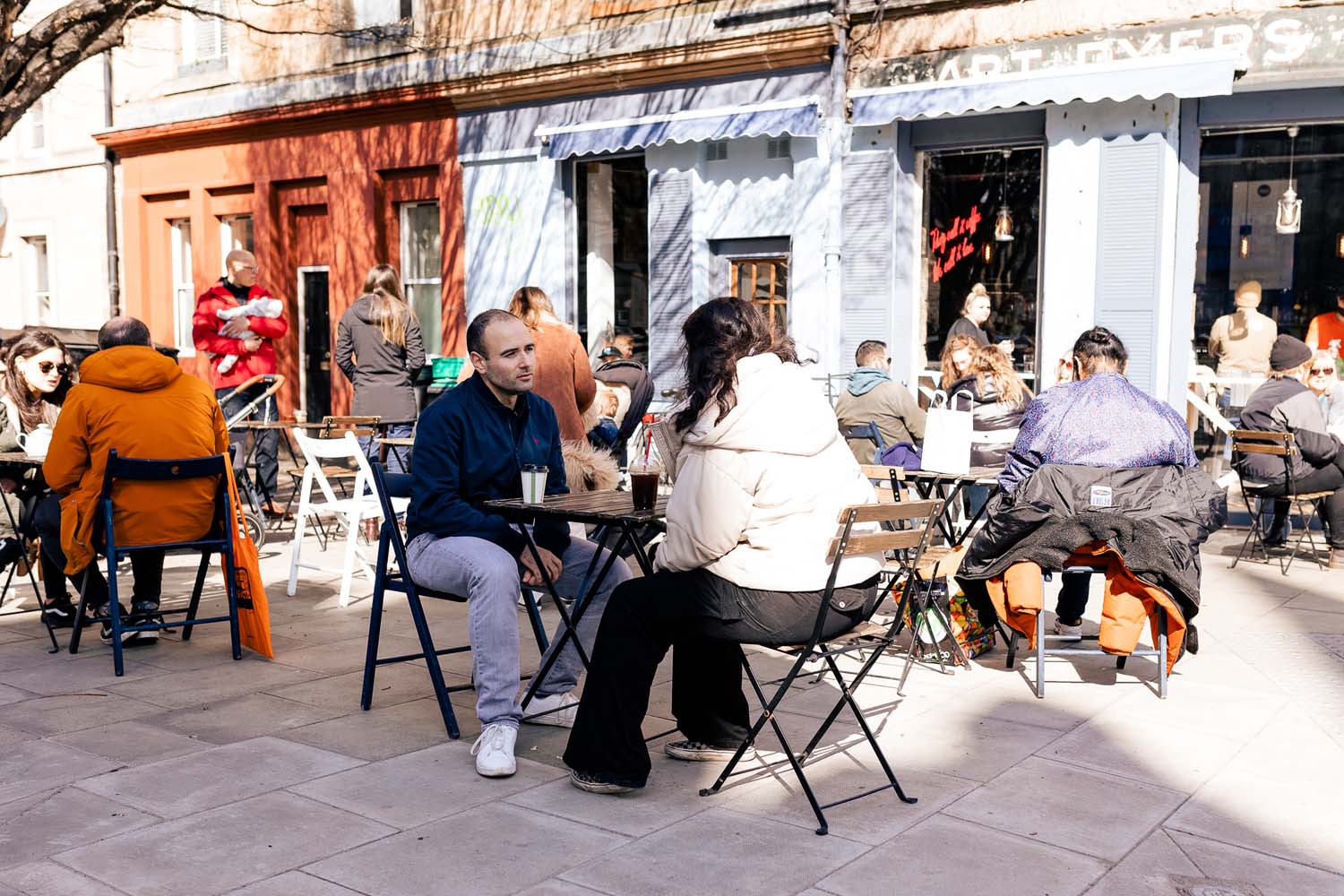 A group of people sitting at small tables outside a cafe on a sunny day 