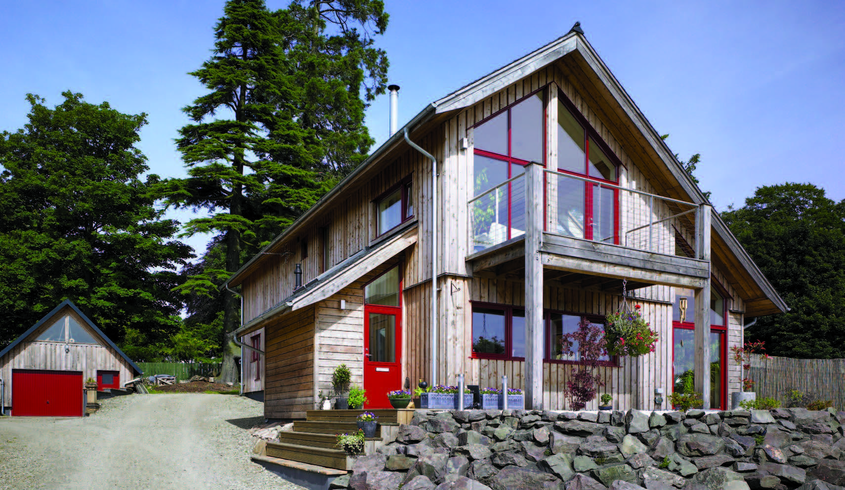 A two-storey house with a vertical timber facade with trees behind it. The doors and window frames are bright red, and the colour of the timber is faded.