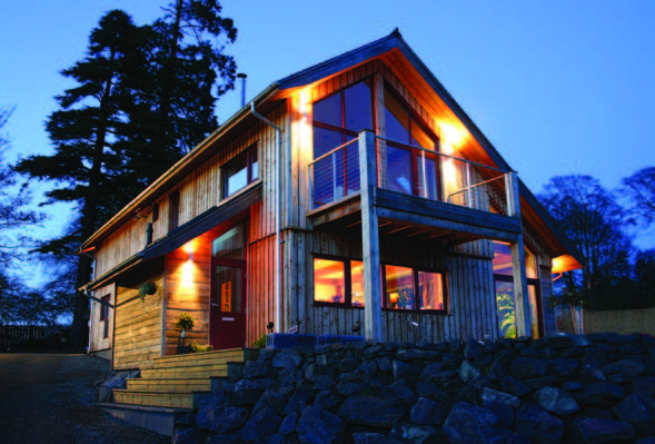 A two-storey house with a vertical timber façade and trees behind it in the evening light. 