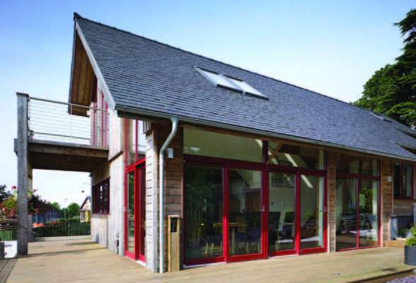 A slate roof with skylights on a longhouse building covered in untreated timber. The side of the house has double sliding red frame doors with windows above it.