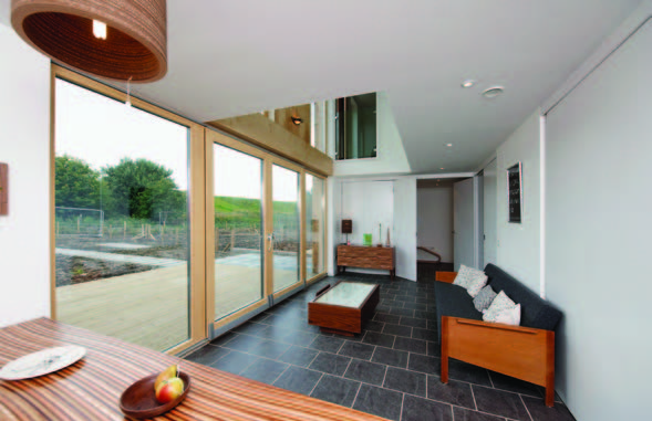 A photograph of the bright interior of a home with large glass windows, wooden window frames and black stone flooring