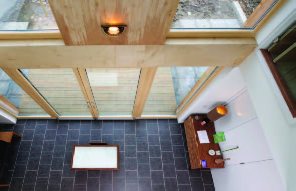 A view from above of a living area with light coloured wooden beams, a black stone floor and large panes of glass.