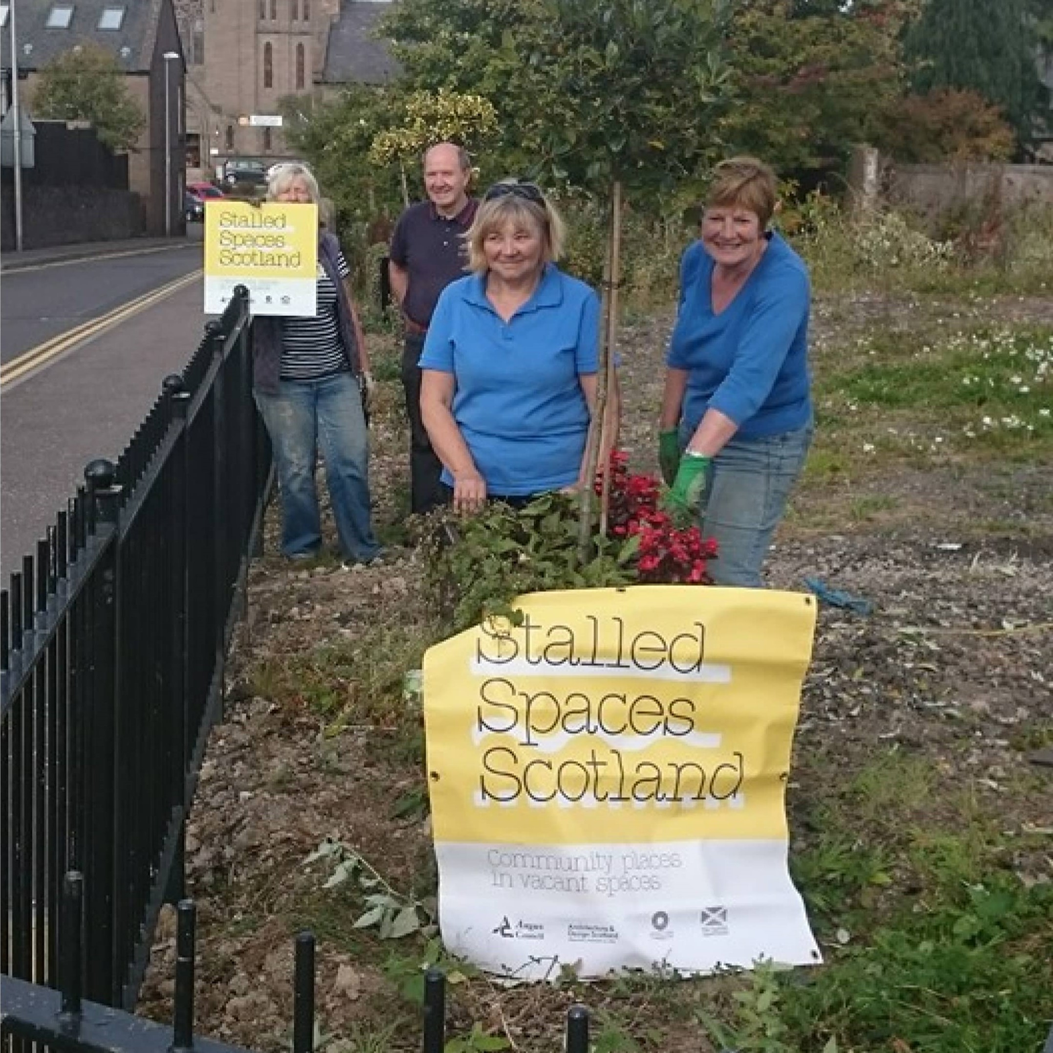 Two women from the Forfar in Flower project hold up a Stalled Spaces Scotland sign in a soon to be green space by a road.