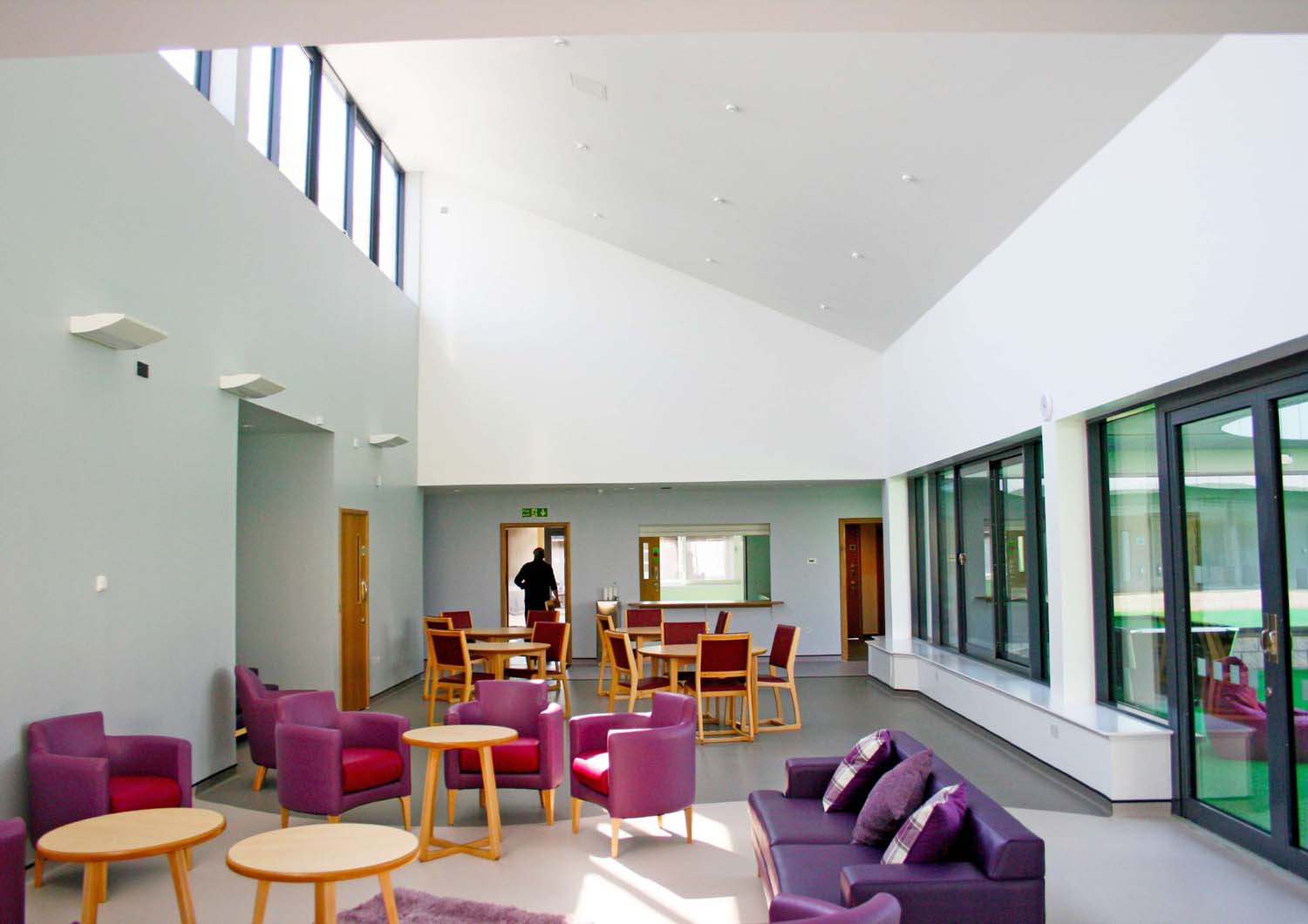 A social space with clerestory windows and a slanted roof. There is a couch and armchairs in purple hues and large windows looking into another interior space.