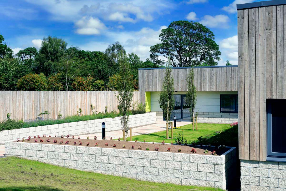 An external view of the timber-cladded entrance of the care unit. There are long raised flowerbeds and a few small trees in front of the entrance, and larger trees in the background..