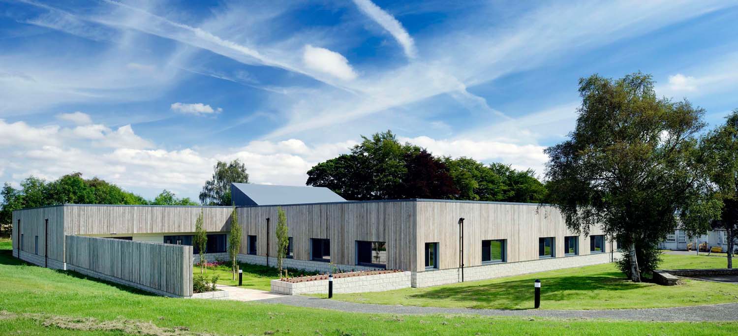 A photograph of a grey timber-clad building sitting into the landscape, against a bright blue sky 