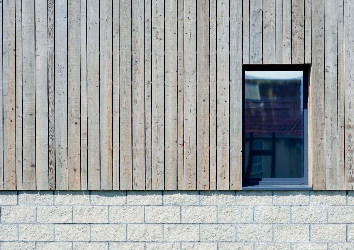 A close up of an exterior wall. There is timber cladding sitting on top of a sandstone base, and a window with a dark frame on the right.