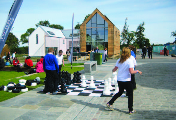 Children playing with a large outdoor chess set at Scotland's Housing Expo with the Flower House in the background.