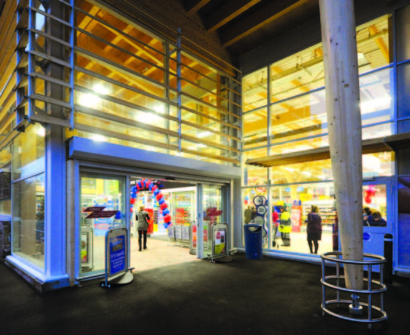   A person walks into the entrance of Tesco Banchory superstore. Timber bladed sunscreens cover the window above the entrance's automatic sliding doors. 