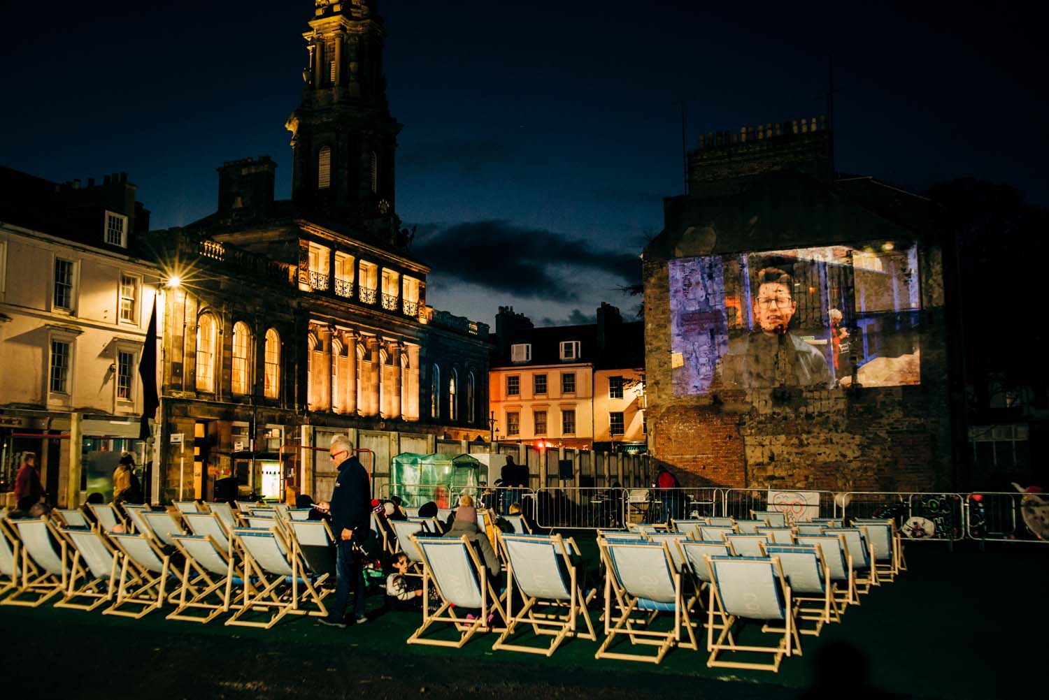 An outdoor film screening in a square with a number of rows of deckchairs and people watching a film projected on a gable end of a stone building