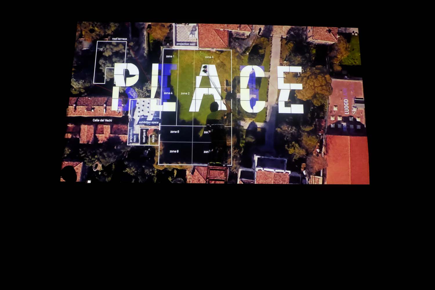 A projection on a wall at nighttime showing the word Place and visible is a man on a ladder 