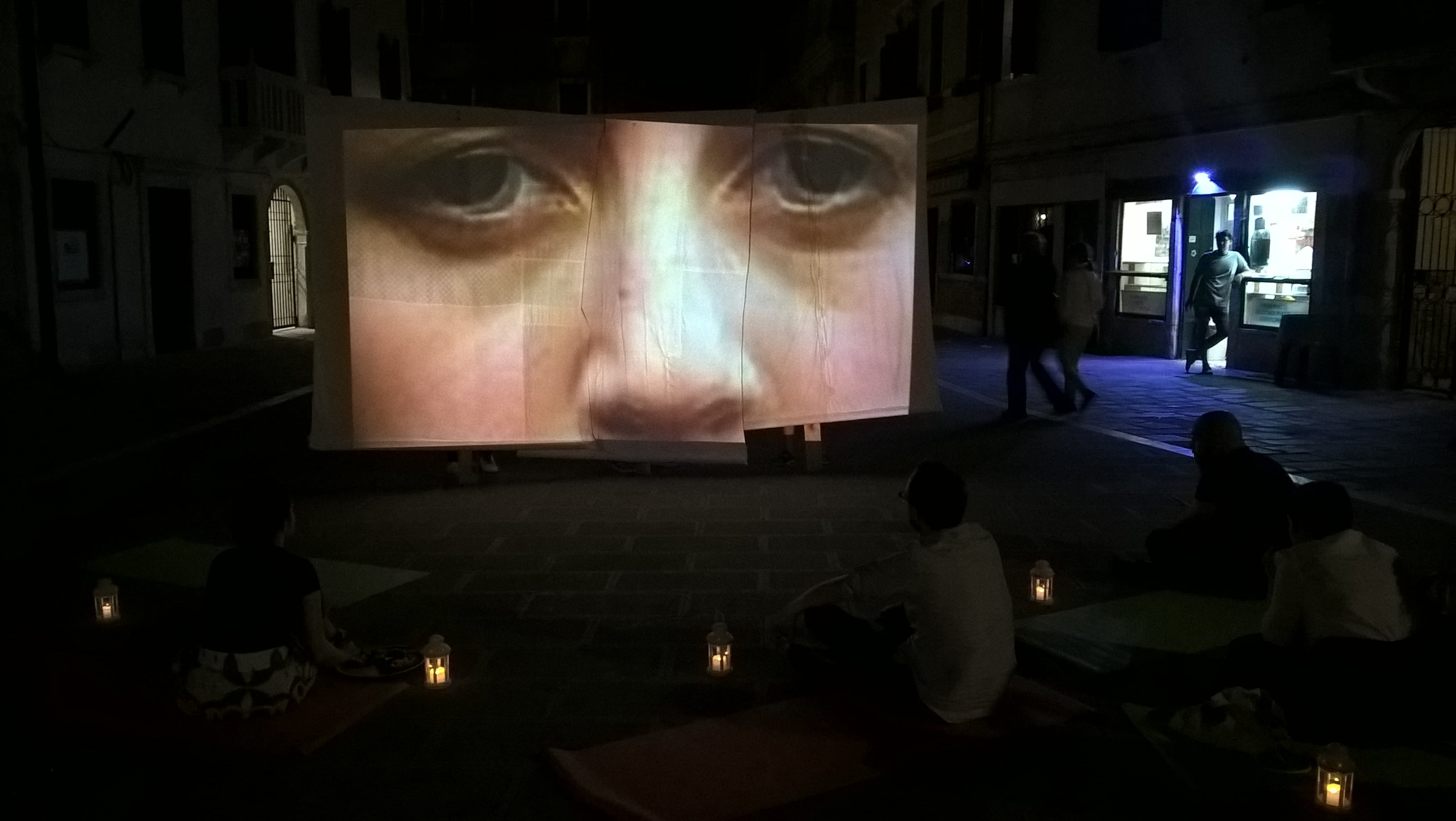 A projection screen in a street is showing a close-up of someone’s face. The scene is dark but candlelit with signs of life.
