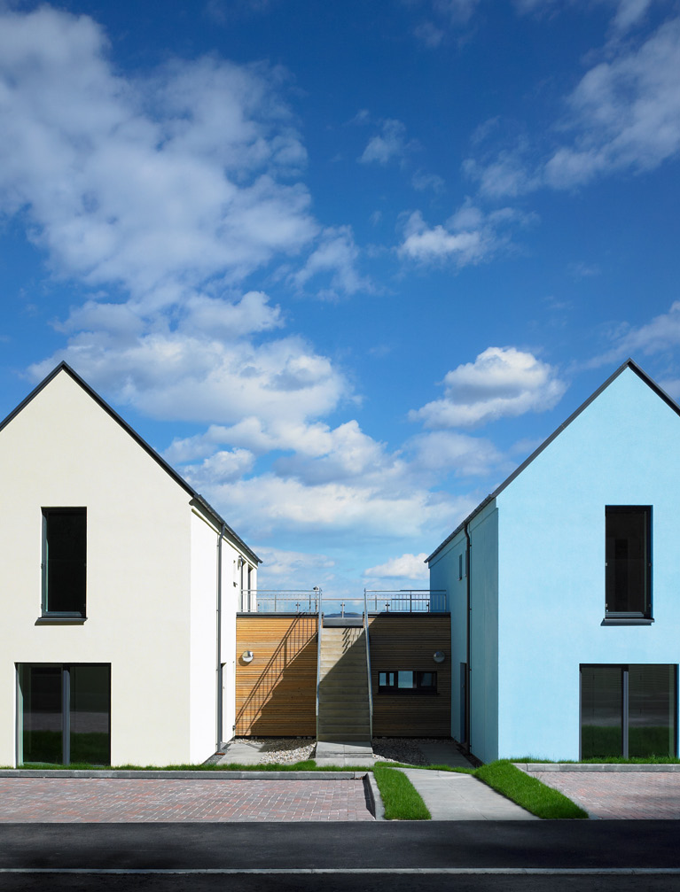 The street facing entrance of two semi-detached houses. The house on the left is white and the right in a light blue tone.