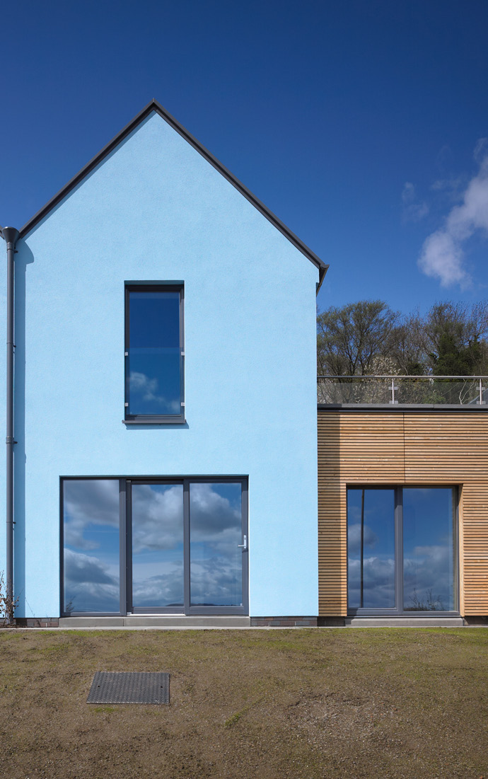 A blue coloured attached house with a sliding doors and a long window. Beside it is a one storey building with vertical wood façade.