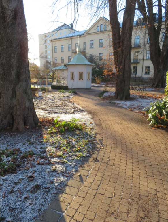 A gently curved brick pathway bends towards a small white painted timber summer house set in a winter garden in front of a large manor house