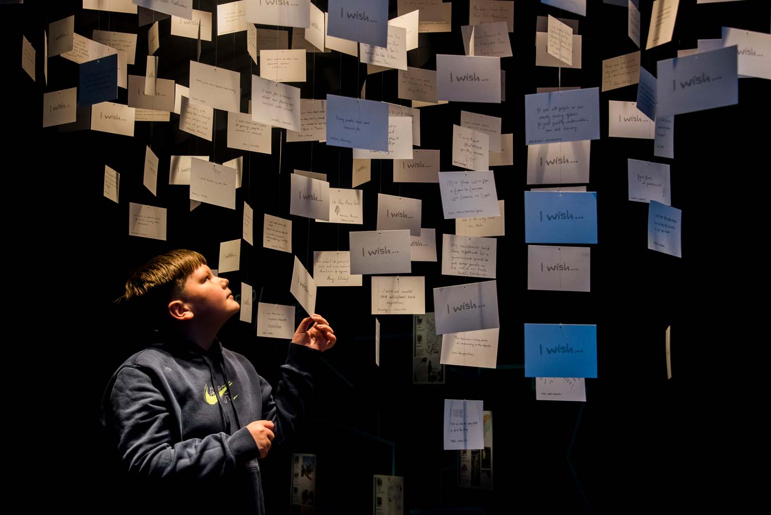 A boy in a dimly lit exhibition space looking up at postcards hanging from the ceiling. The postcards contain wishes of citizens for the future of their place.