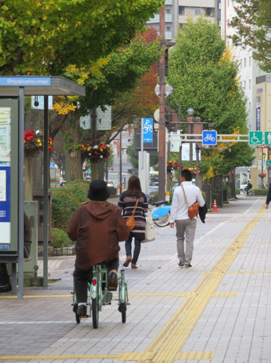 Street scene along a pedestrian friendly road in Toyama, Japan, a cyclist in three wheels rides past a man and woman wearing satchels.