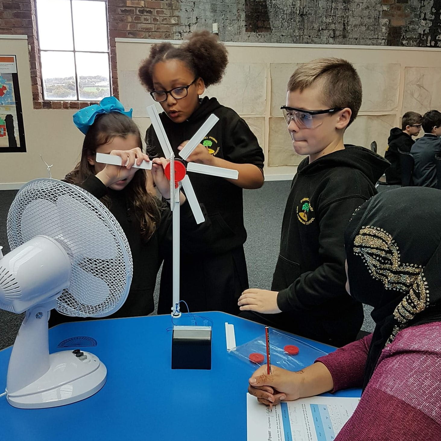 Four children are around a table building a model wind turbine in front of a fan.