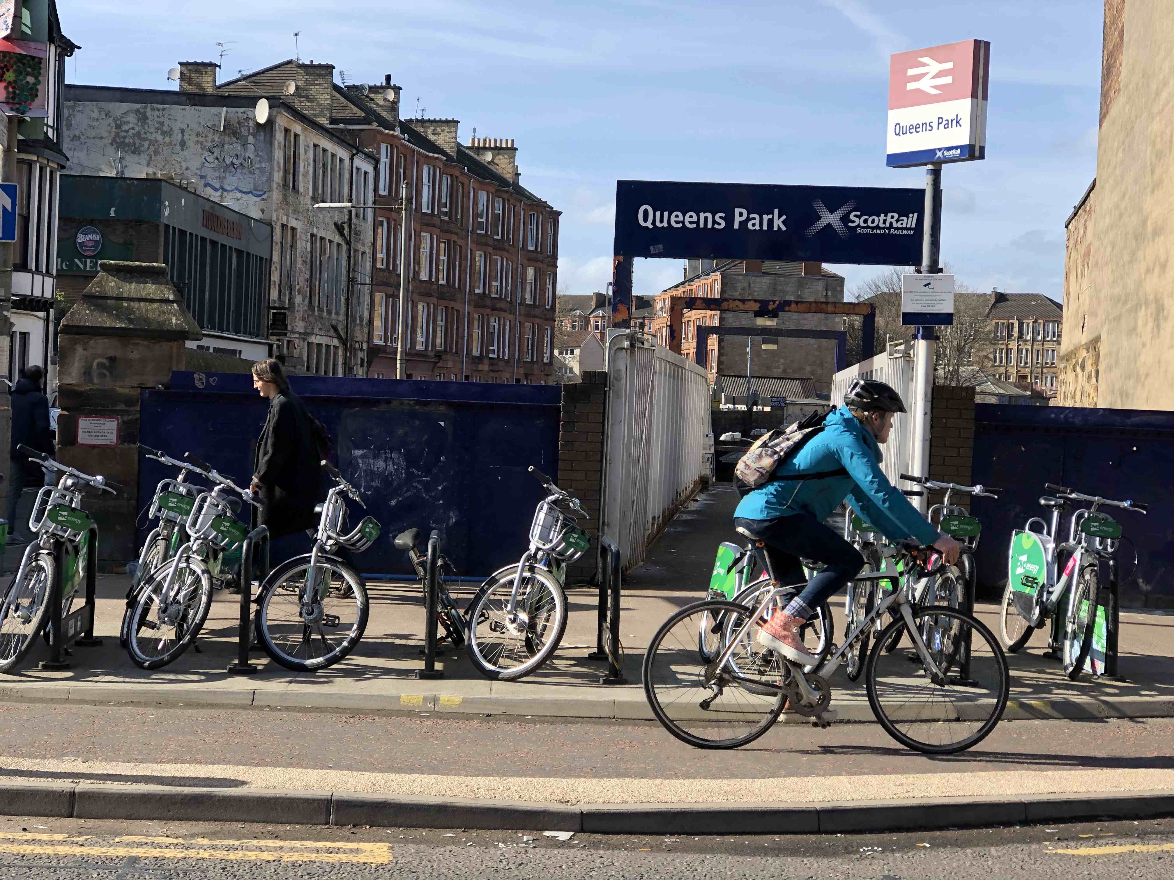 A cyclist is riding past the entrance to Queens Park train station in Glasgow. Someone is walking on the pavement in the opposite direction, and about seven bicycles are parked in a row.