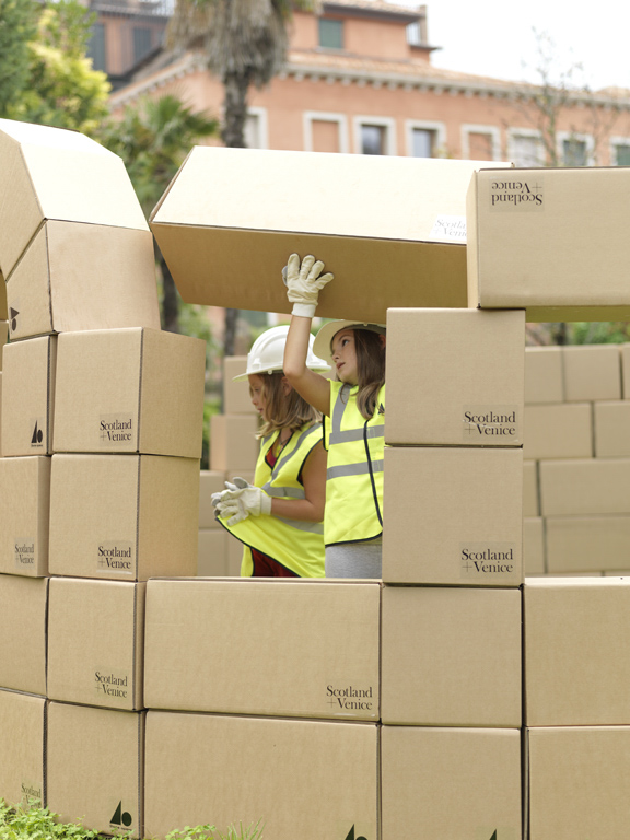 Two young children in high vis jackets and white safety hats are building a wall from large cardboard boxes. Two young children in high vis jackets and white safety hats are building a wall from large cardboard boxes. Two young children in high vis jackets and white safety hats are building a wall from large cardboard boxes.