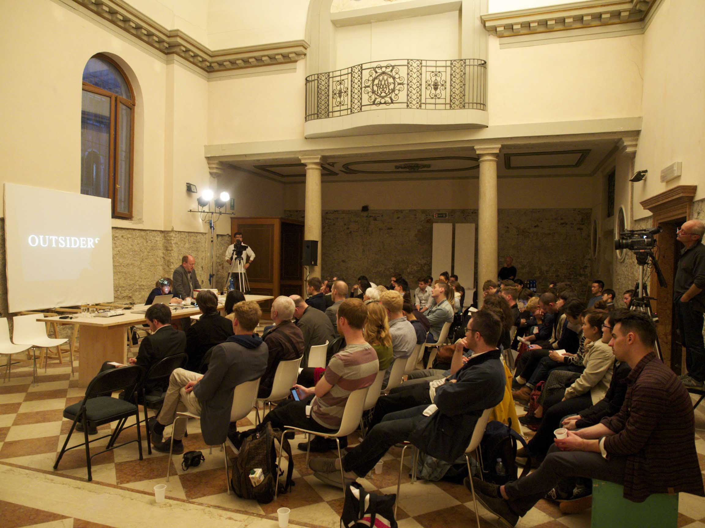 In a large room with two columns and a small balcony, about 50 people are sitting and listening to a presentation. A projector screen displays the word ‘outsider’ in white and all caps.