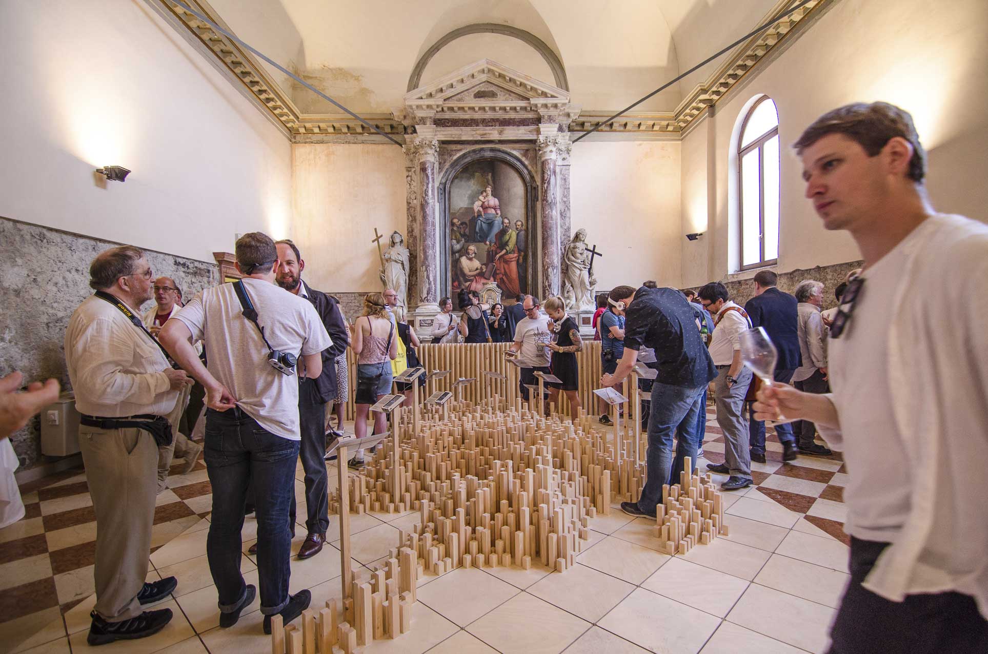 On the floor inside a chapel is a structure in the shape of a dimensional map made from wooden batons of different lengths, each standing upright. Some of the batons hold plaques with mobile phones attached. About 30 people can be seen around the structure.