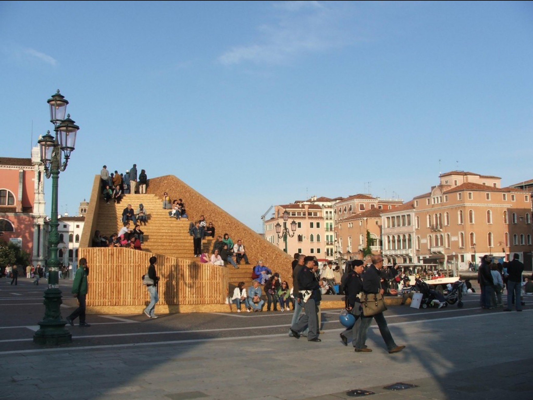 Under a blue sky in a busy Italian piazza is a high wooden structure with steps running up to the top. People are sitting on the steps or looking at the view from the top.