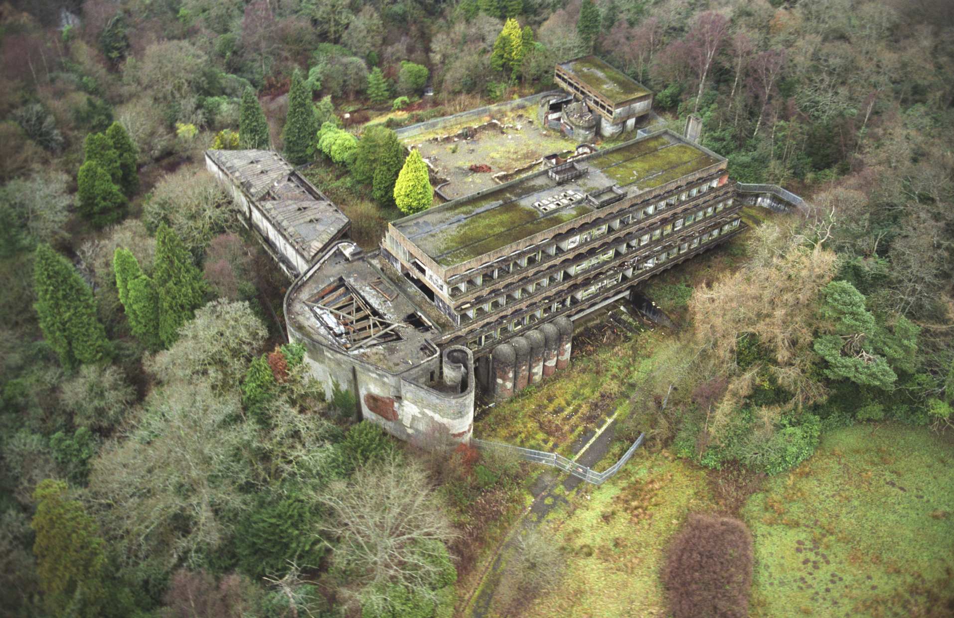 An aerial shot of a large ruin in a wooded area. The ruin is of many different shapes both angled and curved. It is mostly grey with mossy areas.
