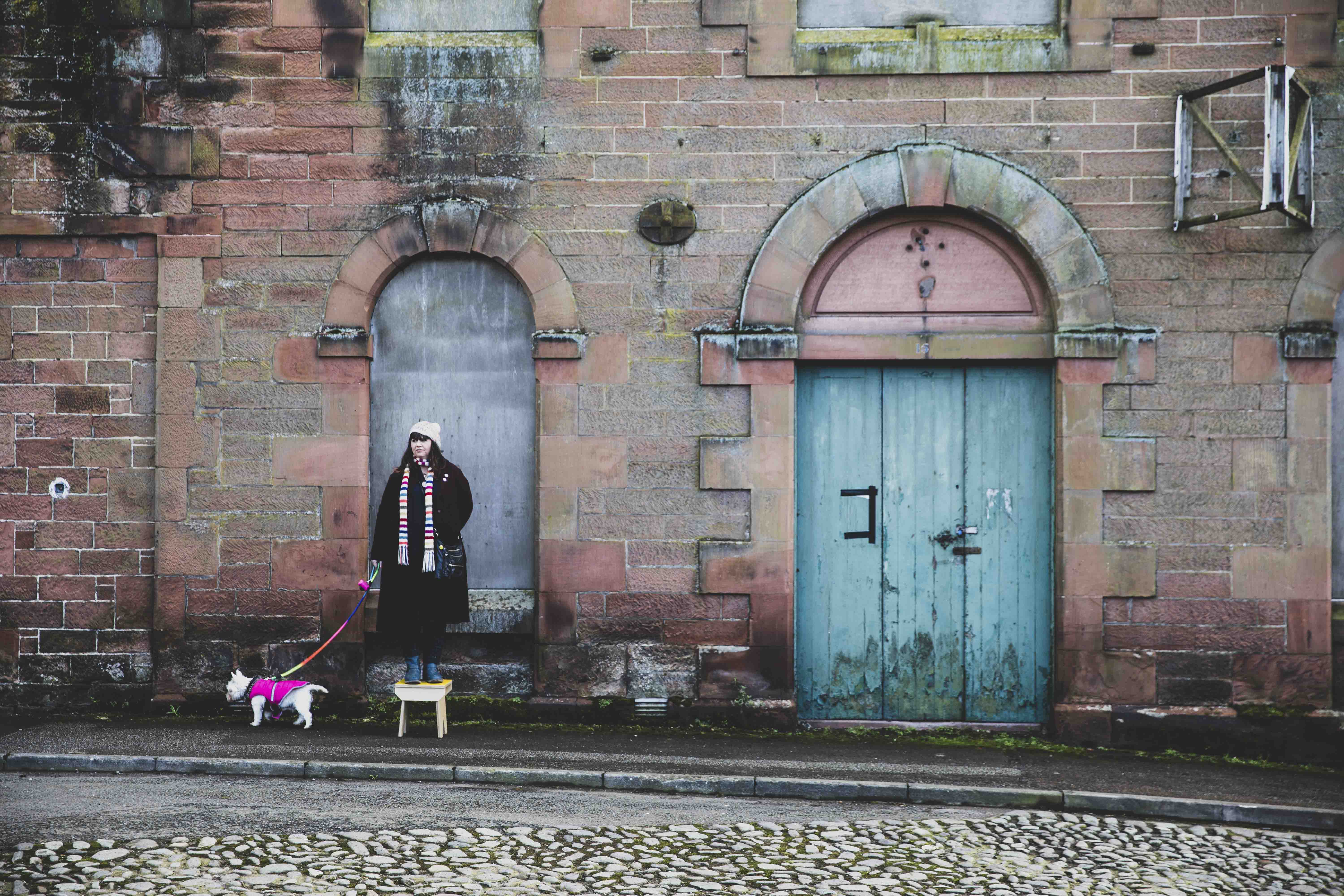 In front of a stained stone wall with blocked up windows and a rotten blue door stands a woman perched on a wooden stool. A small white dog in a bright pink coat is on the street next to her.