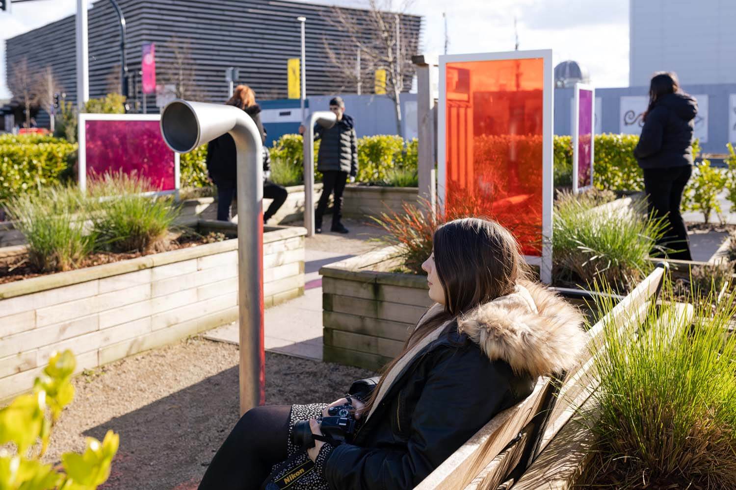 A woman wearing a coat sits on a bench. She is in a garden filled with public artworks.