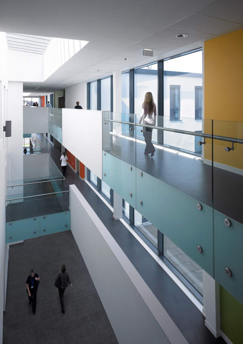 A member of staff walks across the upper circulation space at Renfrew Health and Social Care Centre, featuring floor to ceiling windows and frameless glass balustrades.