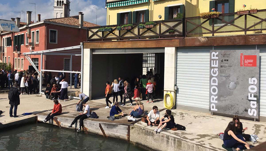 A group of people sit on a canal-side in Venice in front of a yellow building with a large poster