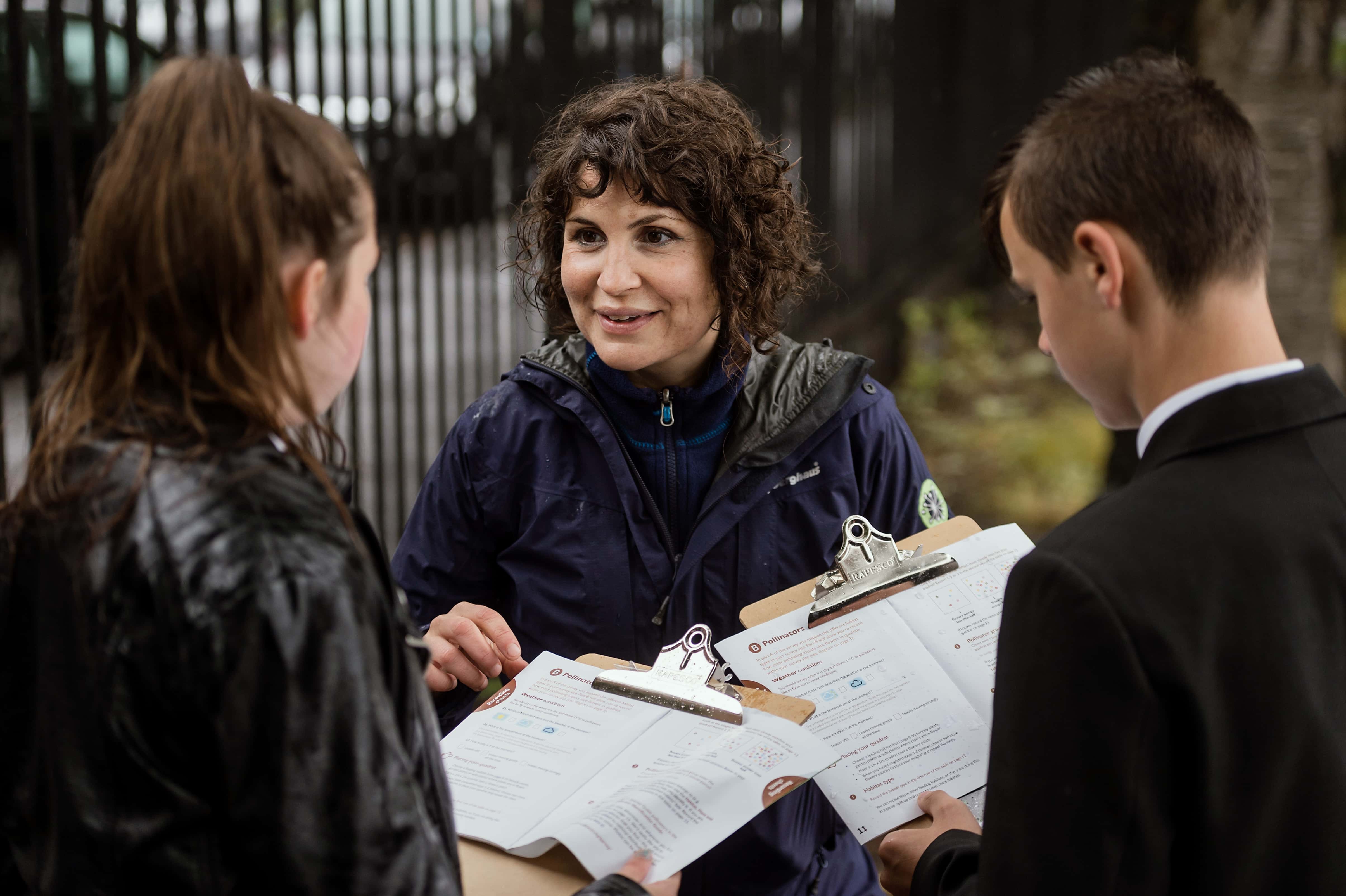 An adult and two teenagers look at checklists on clipboards working together outdoors.