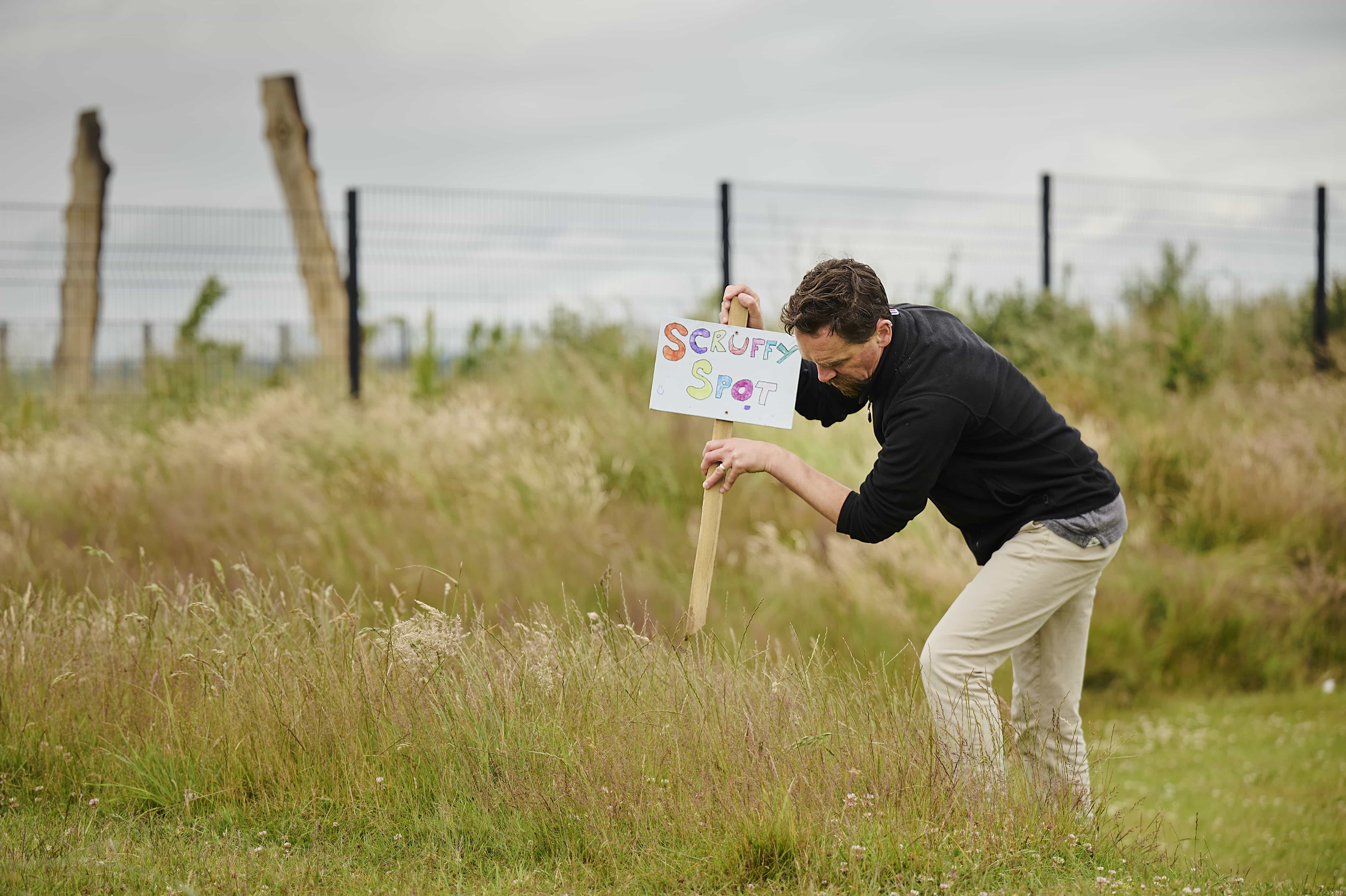 An adult male in a black top and beige trousers plants a sign in long grass. The sign reads 'scruffy area' and is written in crayon.