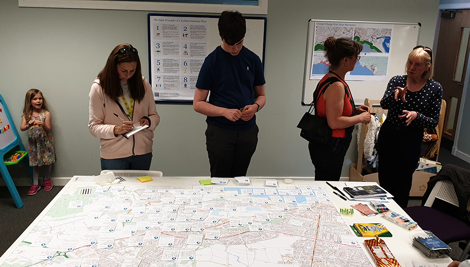 A group of people stand around a table which has a map and a number of sticky notes. A young girls stands in the corner looking at the camera