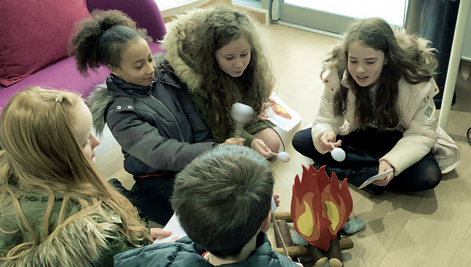 Pupils at Corstorphine primary make s'mores sitting together in a circleabove a fake fire in a room.