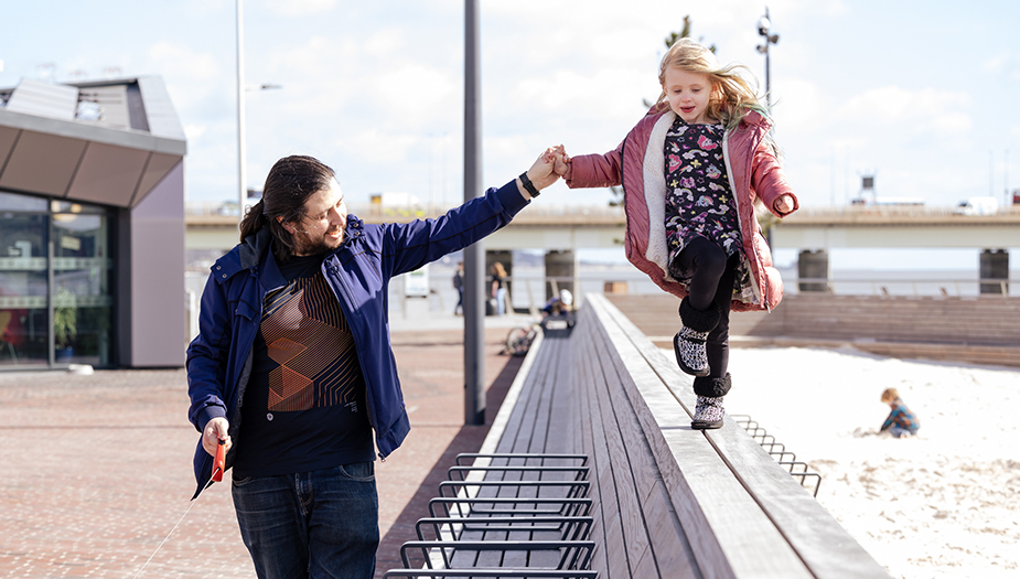 A father and daughter walk across a walled seating bench at Dundee waterfront.