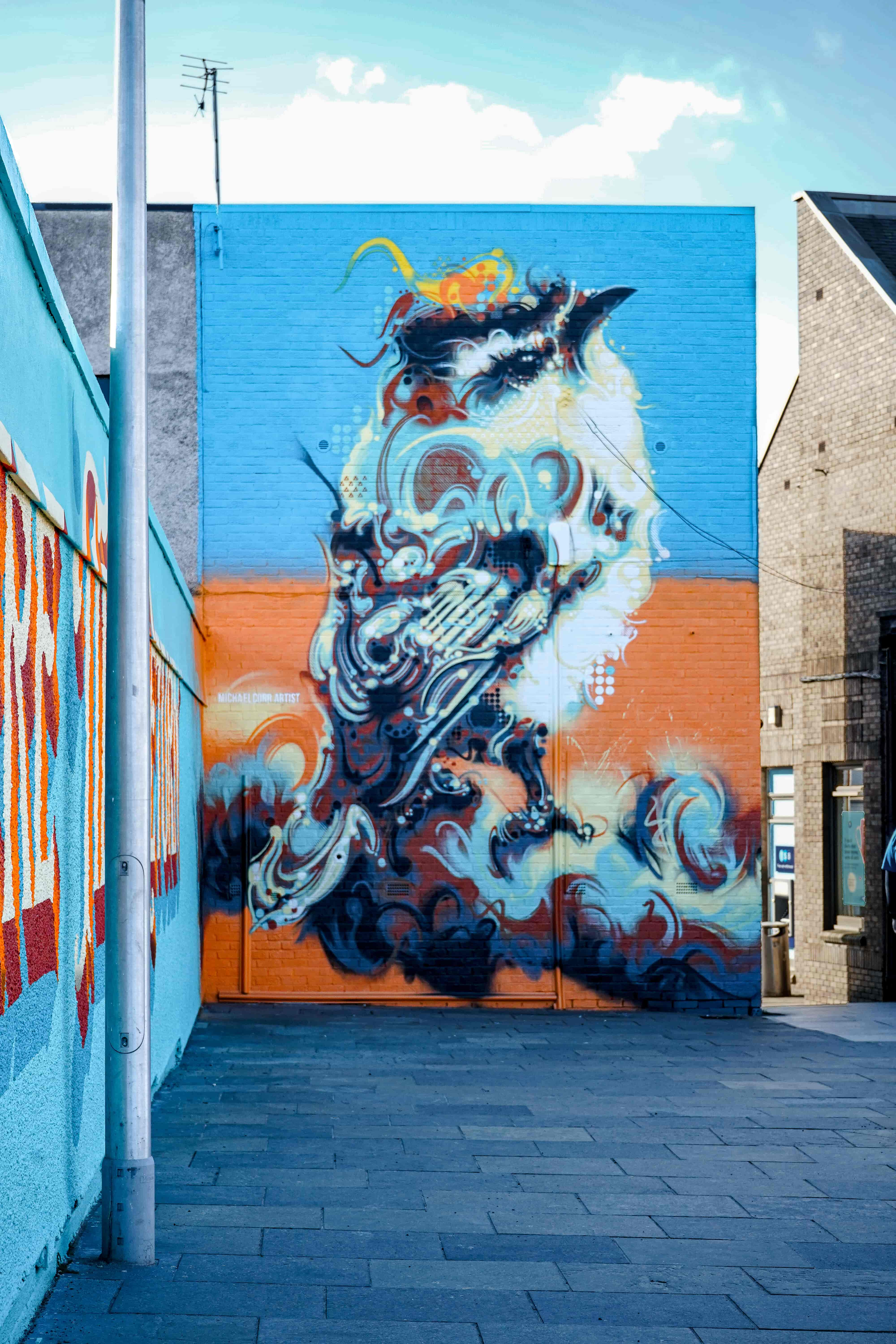 A photograph of a colourful mural on a brick wall. The mural shows a goldcrest bird on a blue and orange background. On the right hand side you can see a gable end of a newly built brick house. 