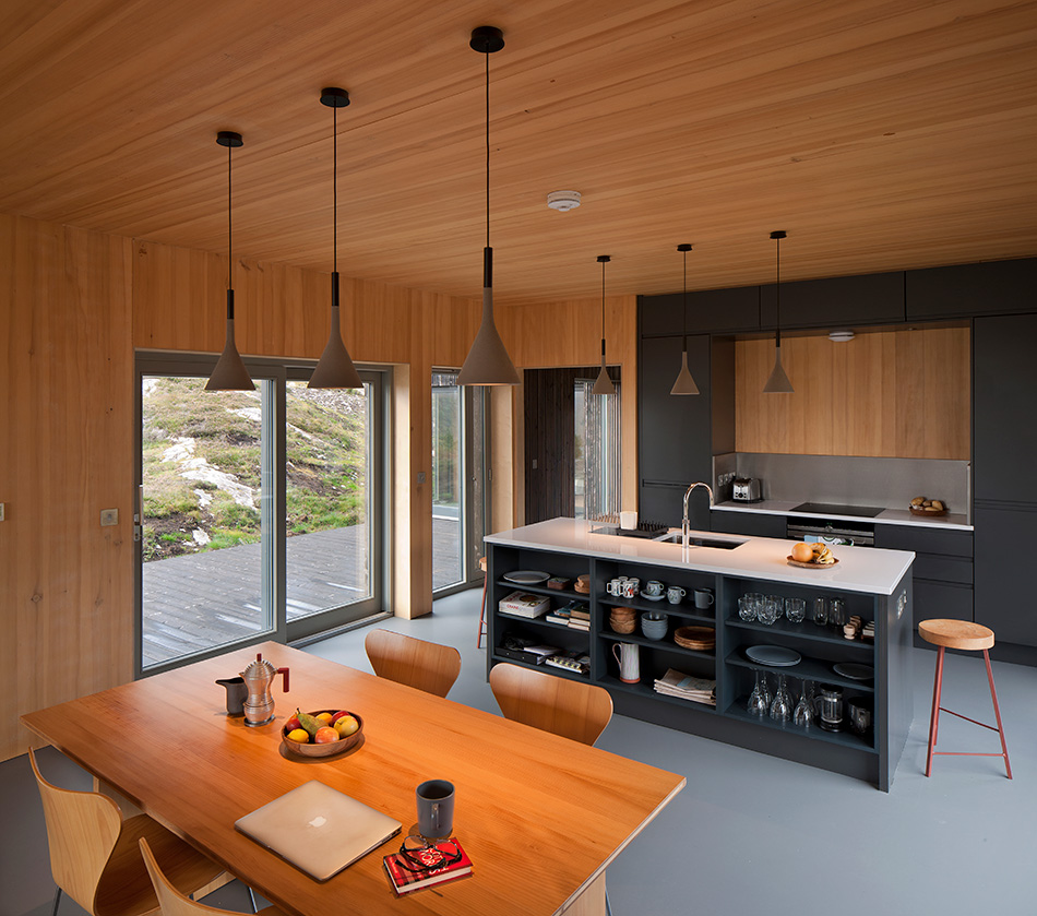The kitchen and dining area from An Cala house. The space is surrounded by interior timber cladding on its walls and ceilings with the space broken up by a walkway between the kitchen and dinging area.