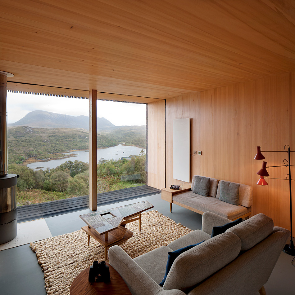 Floor to ceiling windows frame the landscape of An Cala House. A log burner an a infrared heat panel can be seen on either side of the windows.