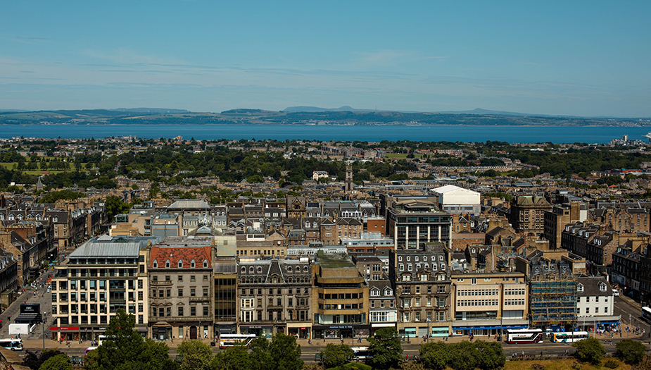 A view of Edinburgh City from the Castle. There is a body of water and mountains in the background.