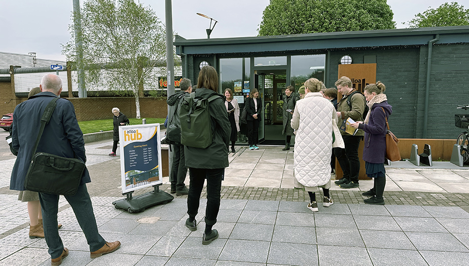 A groiup of people gather around the entrance to a one story darkl grey building - there is a sign at ground level that says Alloa Hub. Nearby there are some other low building and trees in the background.