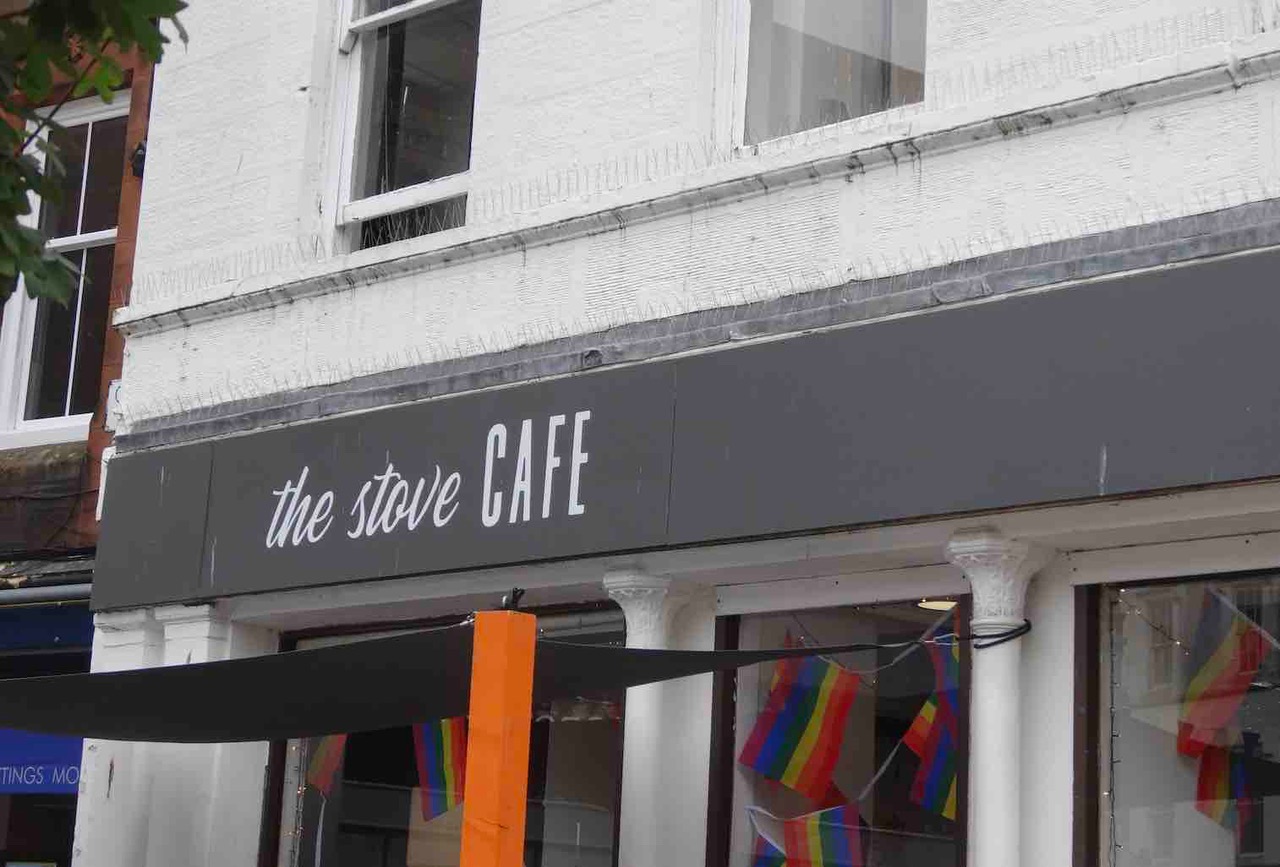 A photograph of a storefront with the words The Stove Cafe written in white on black background. A number of rainbow colour flags are visible in the main window. 