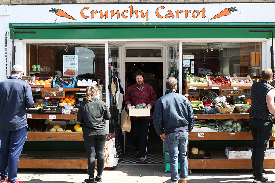 Store front of the Crunchy Carrot with people looking at vegetables and a man walking out with a vegetable box.